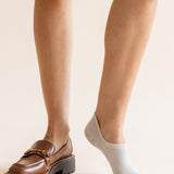 A close-up of a person's legs showing two different styles of low-cut socks: one leg wears a brown leather loafer with a chunky sole, while the other leg wears a light gray ankle sock.