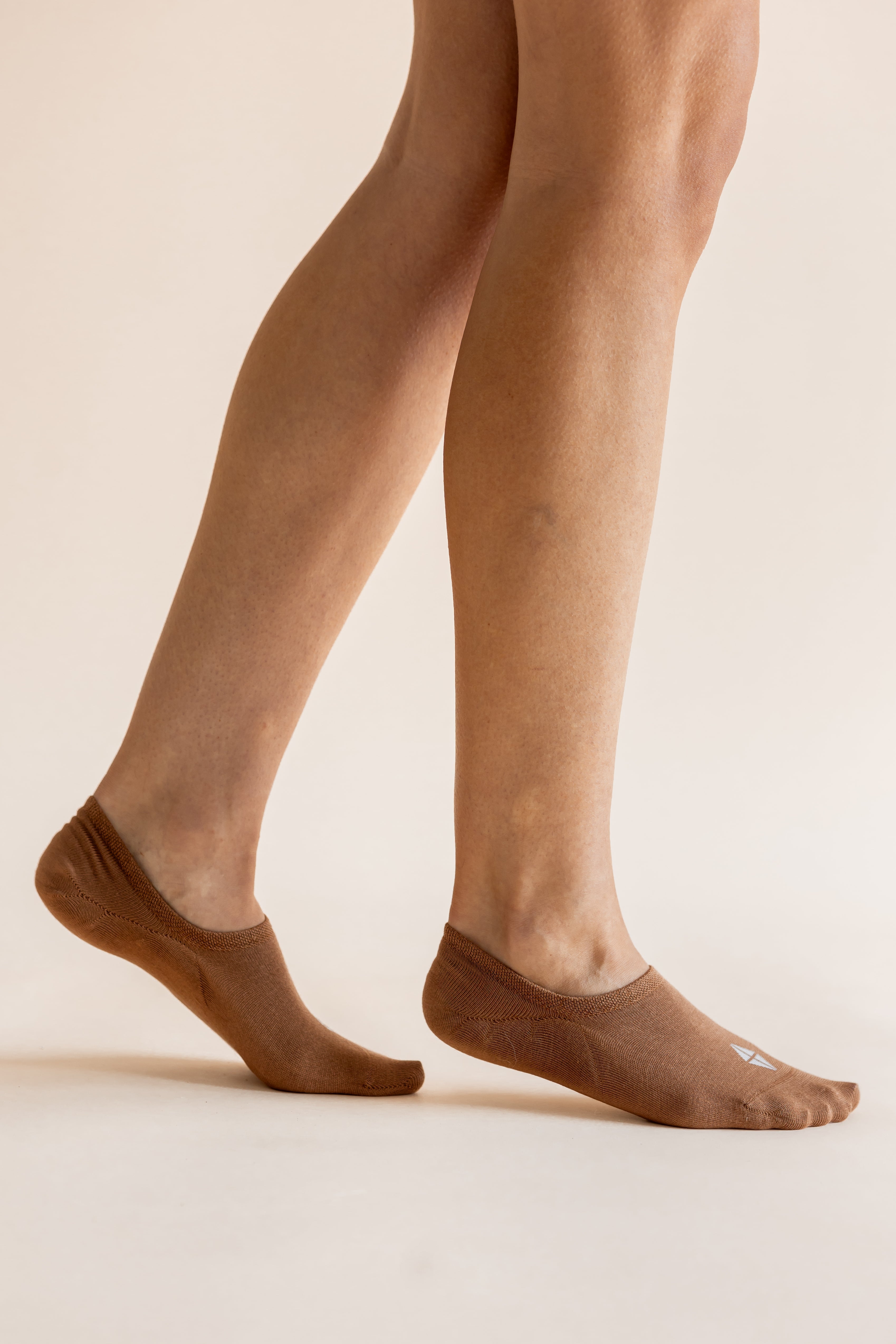 A close-up of a person's legs wearing brown no-show socks against a plain beige studio background.