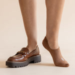 A person wearing brown leather loafers with chunky black platform soles and tan ankle socks, shown from the calves down against a plain beige studio background.
