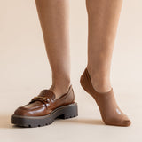 A person wearing brown leather loafers with chunky black platform soles and tan ankle socks, shown from the calves down against a plain beige studio background.