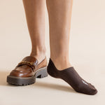 A person wearing dark brown ankle socks and brown leather loafers with chunky black soles, standing against a plain beige studio background.