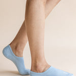 A close-up of a person's legs wearing light blue low-cut no-show socks against a solid beige background.