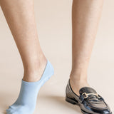 A close-up of a person's legs showing one ankle wearing a light blue no-show sock and the other wearing a black leather loafer with a gold buckle detail, set against a solid beige background.