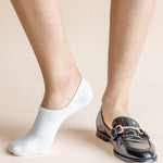 A close-up of a person's feet wearing a white low-cut ankle sock on one foot and a black patent leather loafer with a gold-tone buckle on the other, set against a solid light beige background.