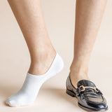 A close-up of a person's feet wearing a white low-cut ankle sock on one foot and a black patent leather loafer with a gold-tone buckle on the other, set against a solid light beige background.