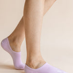A close-up of a person's feet wearing light lavender low-cut no-show socks against a solid pale beige background.