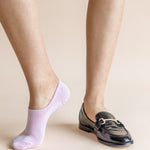 A person's legs shown from the calves down, wearing a light lavender no-show sock on one foot and a black patent leather loafer with a gold buckle on the other, against a solid beige background.