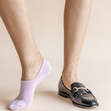 A person's legs shown from the calves down, wearing a light lavender no-show sock on one foot and a black patent leather loafer with a gold buckle on the other, against a solid beige background.