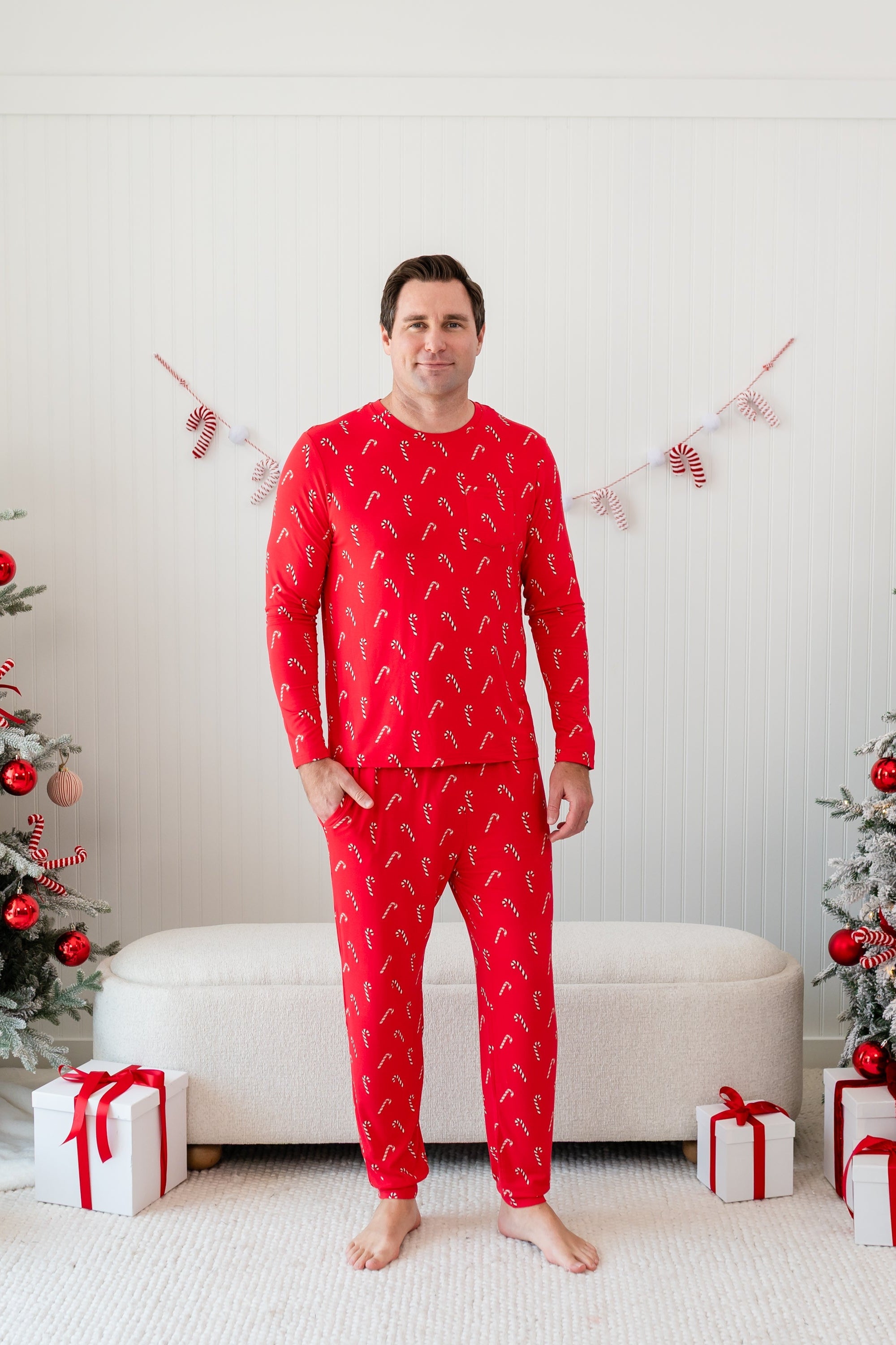 Male model standing in front of a cream ottoman between two decorated Christmas trees wearing the Men's Jogger Set in Candy Cane with one hand in the jogger pants pocket