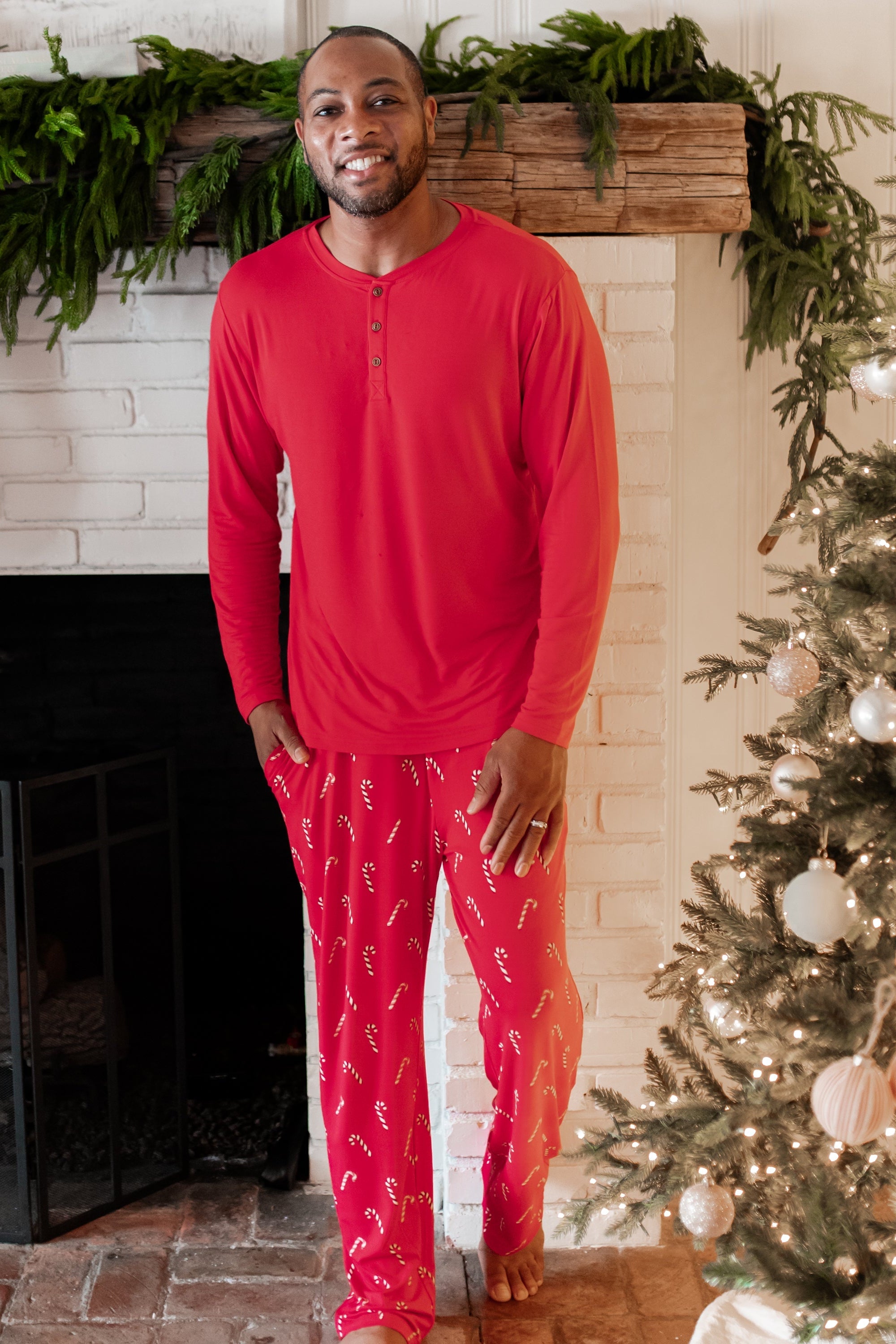 Male model standing in front of a white brick fireplace wearing the Men's Lounge Pants in Candy Cane and Men's Henley top in Cardinal