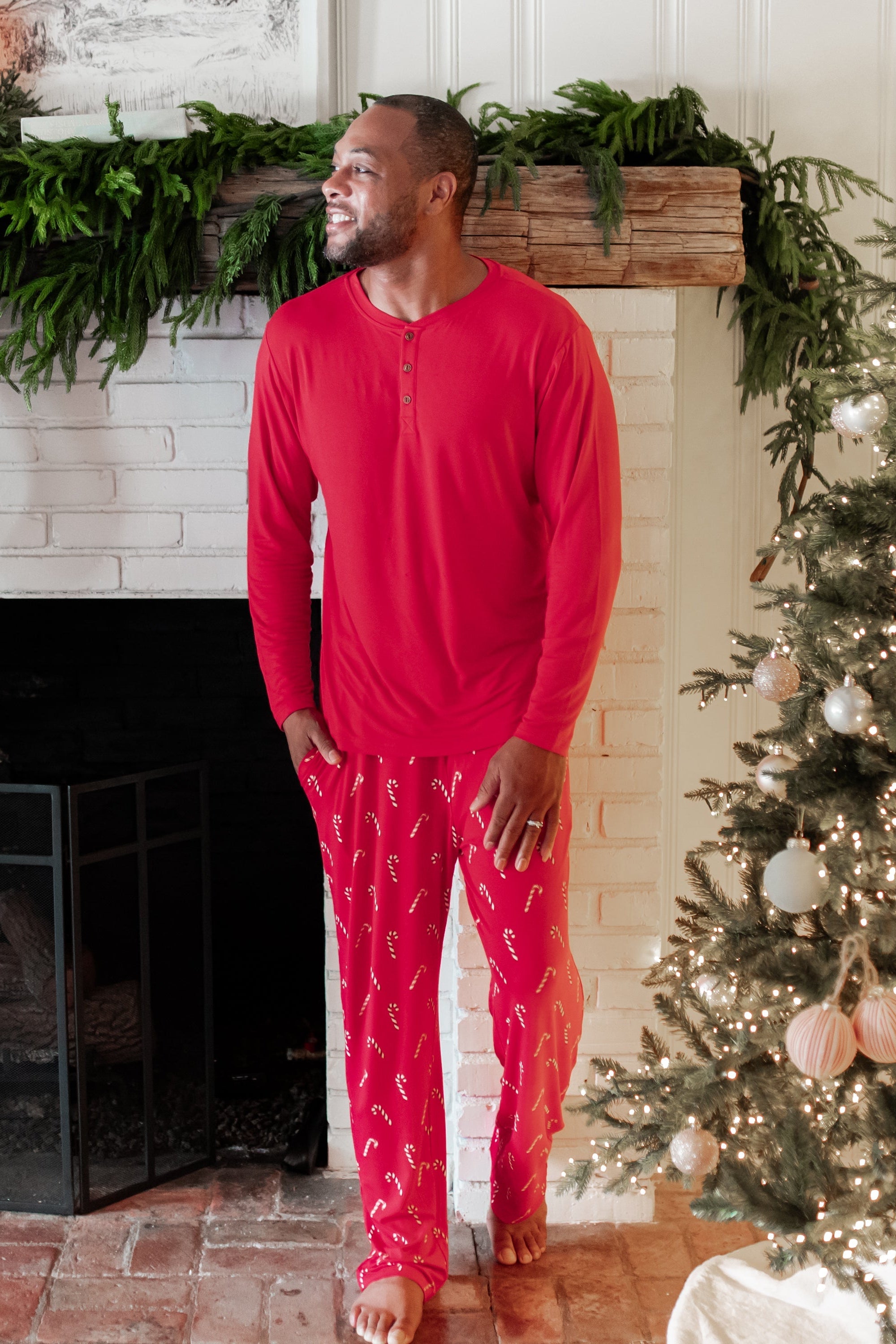 Male model walking looking off to the side in front of a white brick fireplace wearing the Men's Lounge Pants in Candy Cane and Cardinal men's Henley top