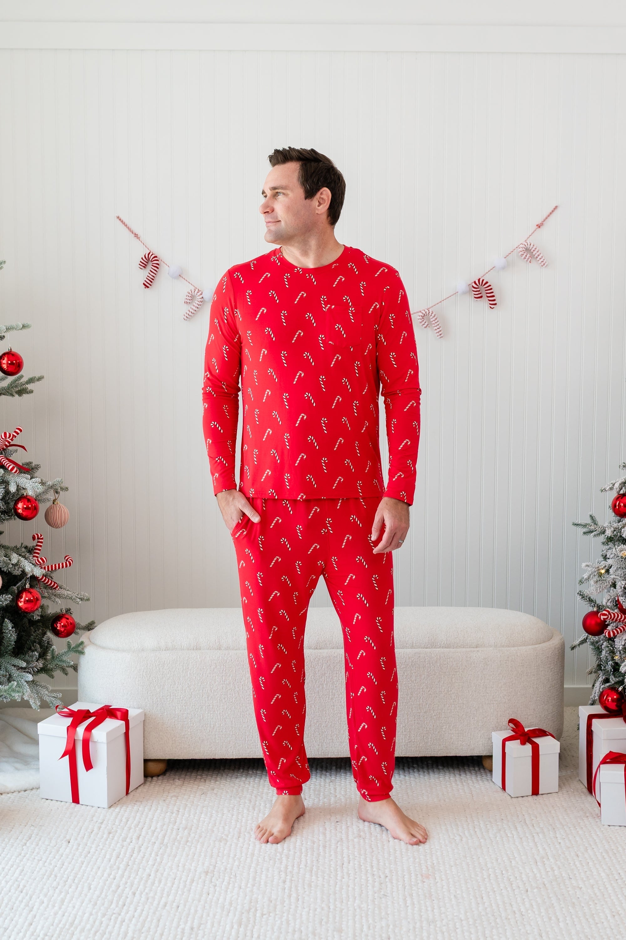 Male model standing with one hand in his pocket wearing the Men's Jogger Set in Candy Cane in front of a cream ottoman between two decorated Christmas trees