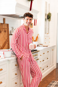Modal wearing Men's Long Sleeve Pajama Set in red and white Peppermint Stripes print standing in a kitchen while holding a cup
