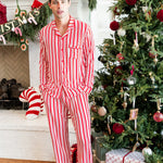 Modal wearing Men's Long Sleeve Pajama Set in red and white Peppermint Stripes print with Christmas tree and present in the background