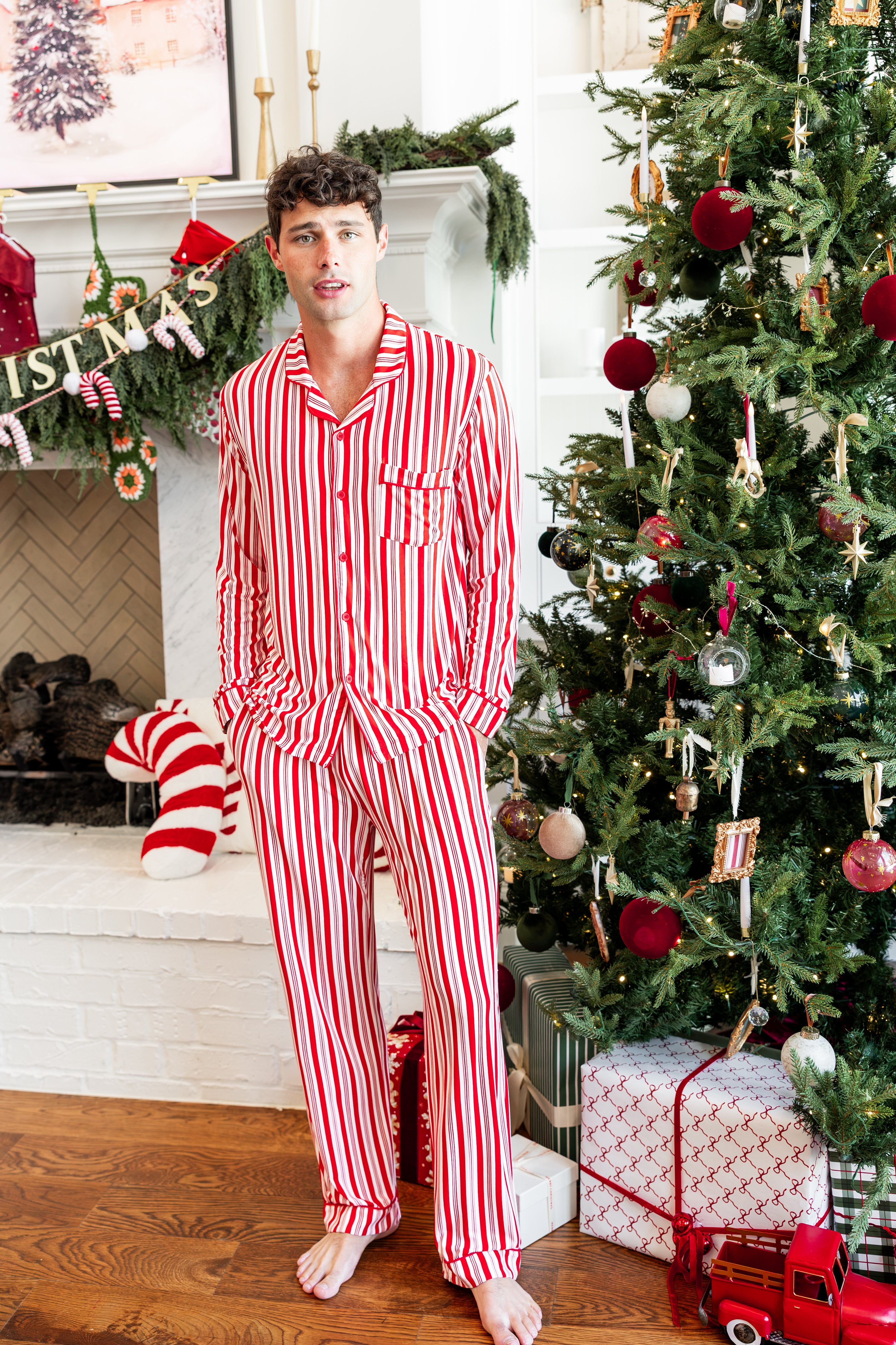 Modal wearing Men's Long Sleeve Pajama Set in red and white Peppermint Stripes print with Christmas tree and present in the background