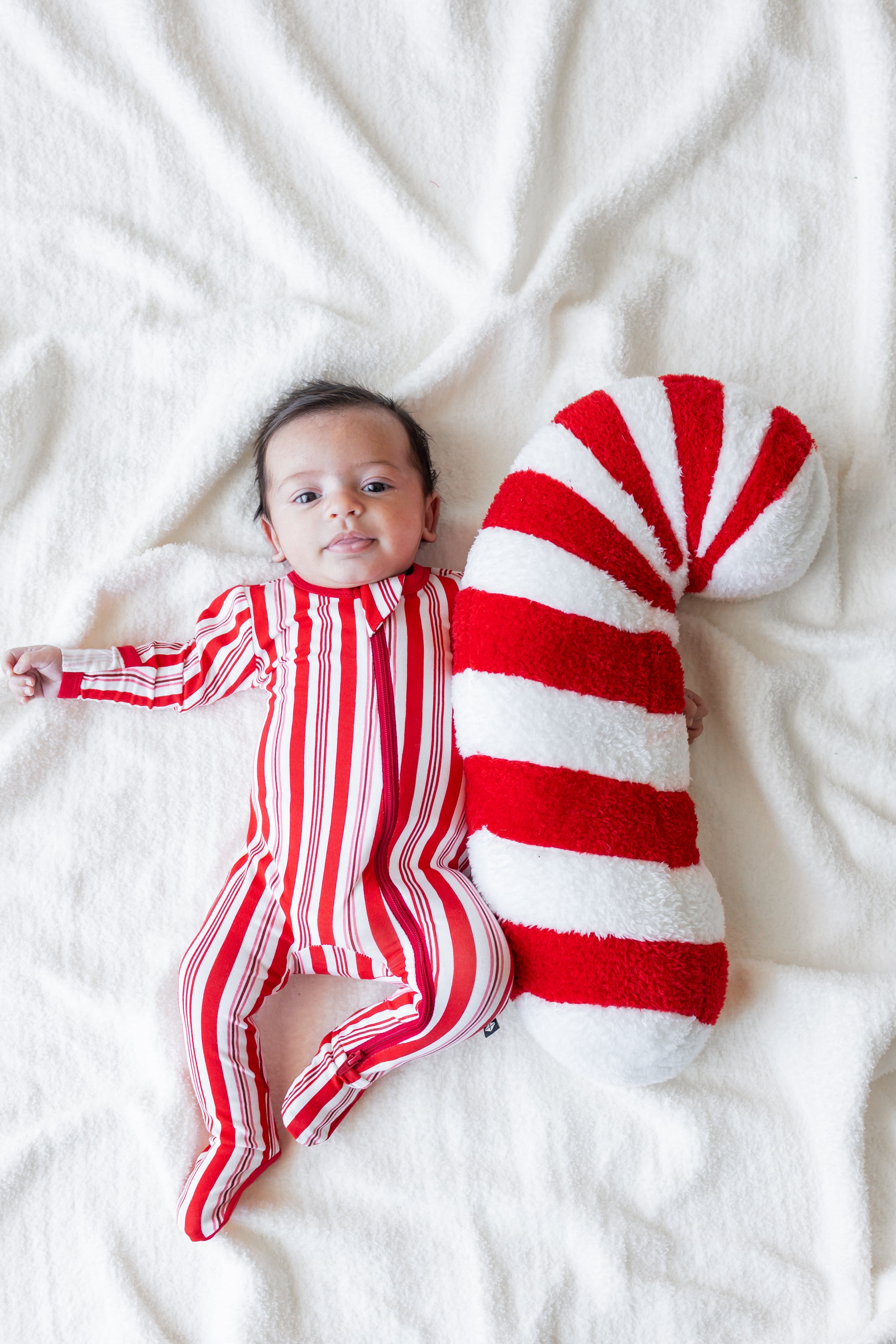 Baby wearing Zippered Footie in red and white Peppermint Stripes print lying with a candy cane prop on a white blanket