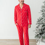 Smiling model standing in front of a white paneled wall and next to a Christmas tree while wearing the Men's Long Sleeve Pajama Set in Candy Cane