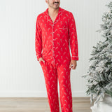 Smiling model standing in front of a white paneled wall and next to a Christmas tree while wearing the Men's Long Sleeve Pajama Set in Candy Cane