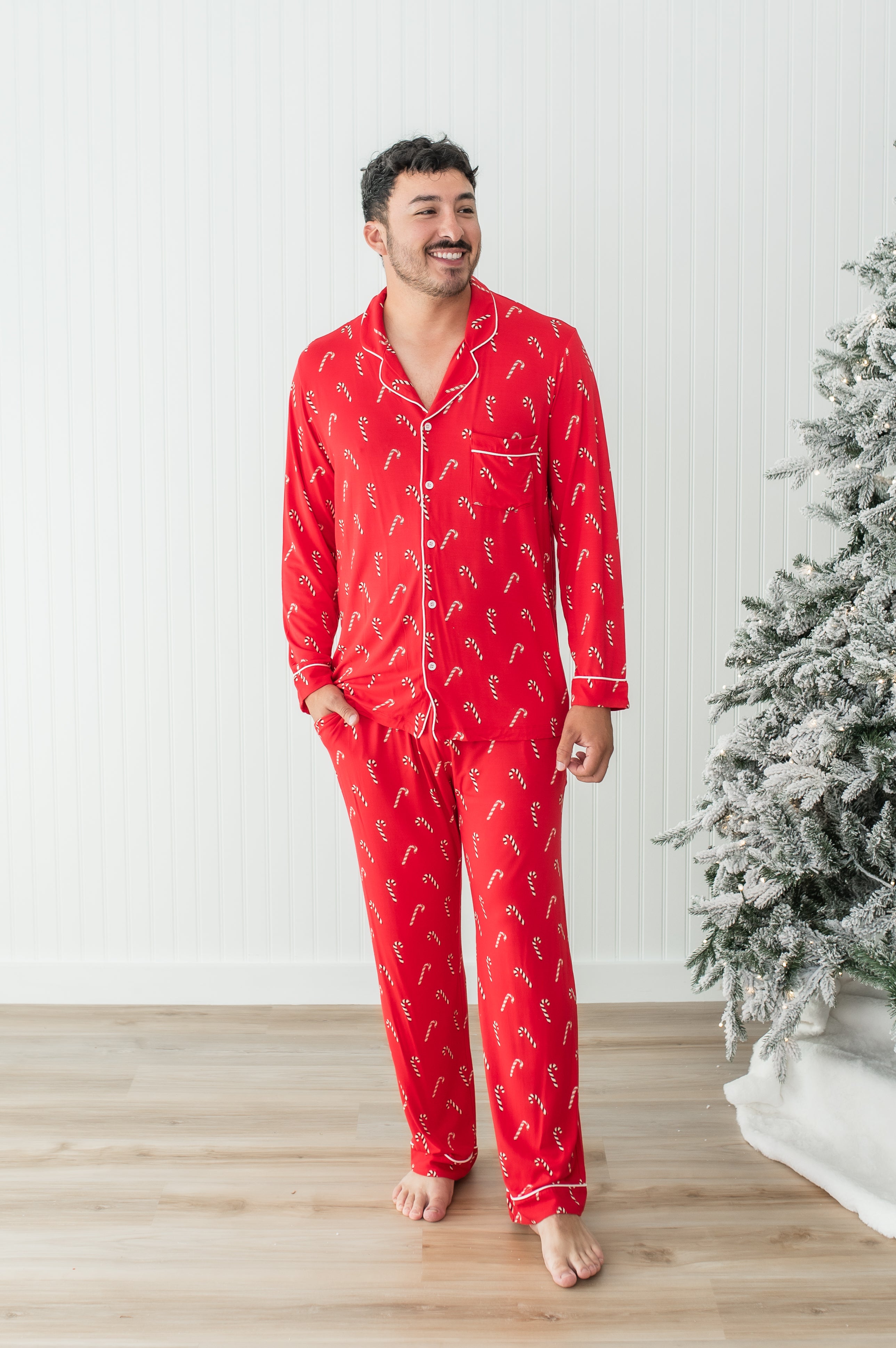 Smiling model standing in front of a white paneled wall and next to a Christmas tree while wearing the Men's Long Sleeve Pajama Set in Candy Cane