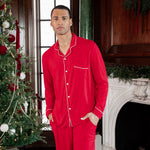 A man wearing a bright red long-sleeve pajama set with white piping stands in a decorated holiday room with a Christmas tree and fireplace.