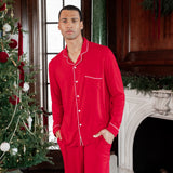 A man wearing a bright red long-sleeve pajama set with white piping stands in a decorated holiday room with a Christmas tree and fireplace.