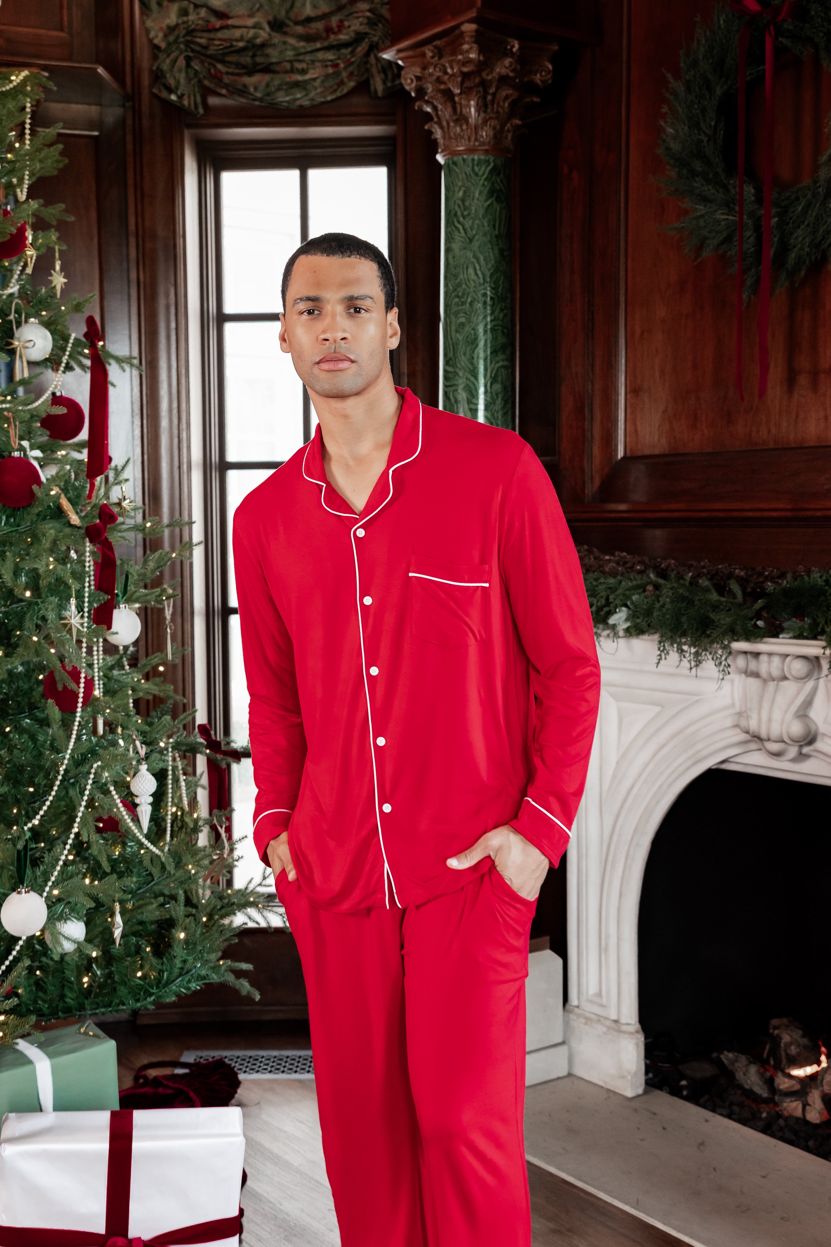A man wearing a bright red long-sleeve pajama set with white piping stands in a decorated holiday room with a Christmas tree and fireplace.