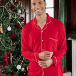 A man wearing a bright red long-sleeve pajama set with white piping stands in front of a decorated Christmas tree in an indoor holiday setting.