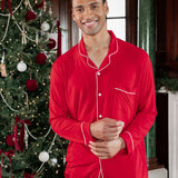 A man wearing a bright red long-sleeve pajama set with white piping stands in front of a decorated Christmas tree in an indoor holiday setting.