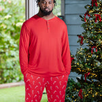 Model standing next to Christmas tree while modeling the Men's Long Sleeve Henley Top in Cardinal paired with Lounge Pants in Candy Cane