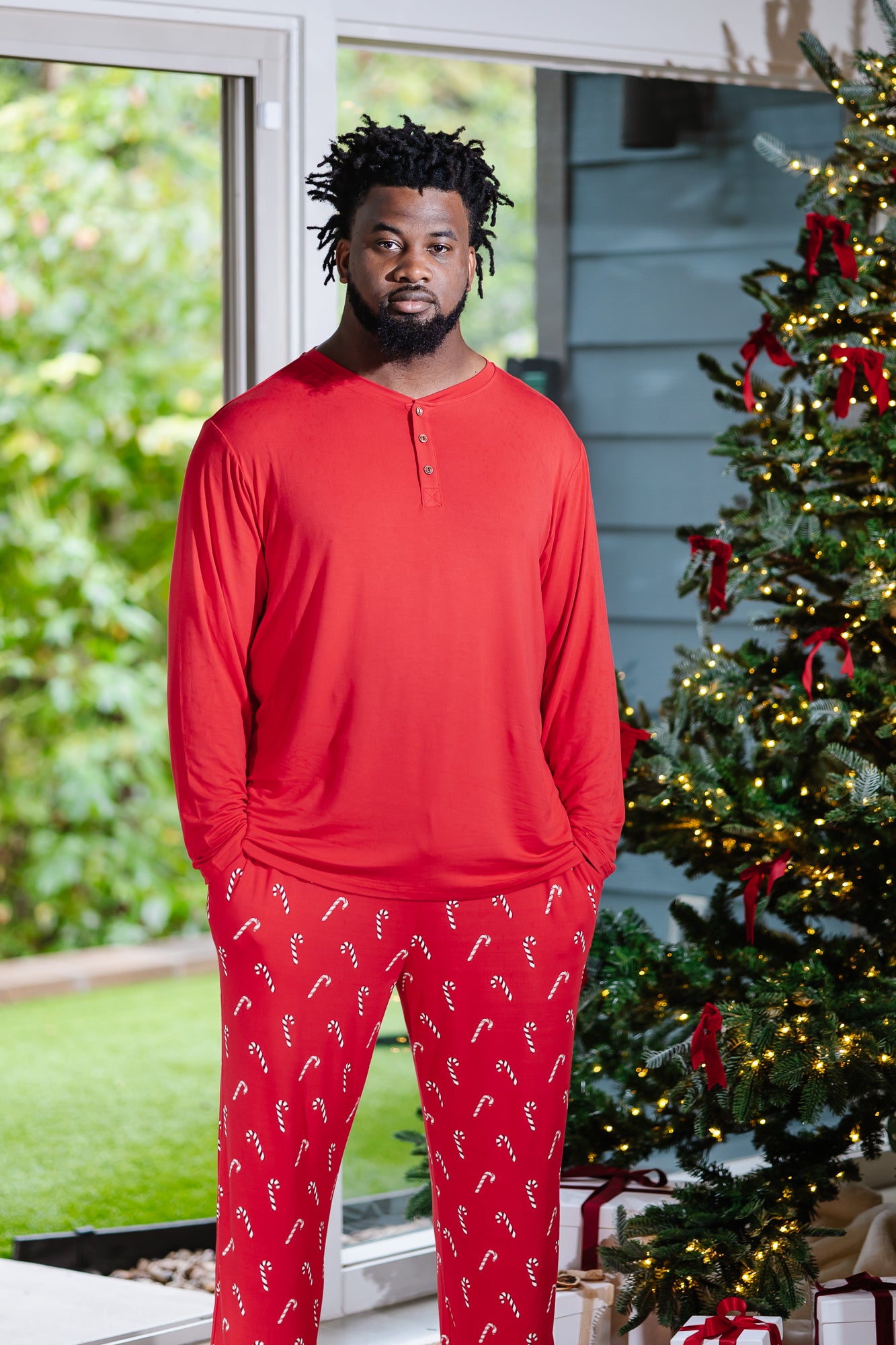 Model standing next to Christmas tree while modeling the Men's Long Sleeve Henley Top in Cardinal paired with Lounge Pants in Candy Cane