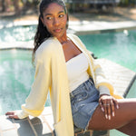 Female model sitting pool side wearing the Chunky Knit Women's Oversized Cardigan in Chamomile overtop a white shirt and medium wash jean shorts