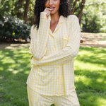 A woman wearing a light yellow and white gingham pajama set stands outdoors on a grassy lawn with trees in the background.