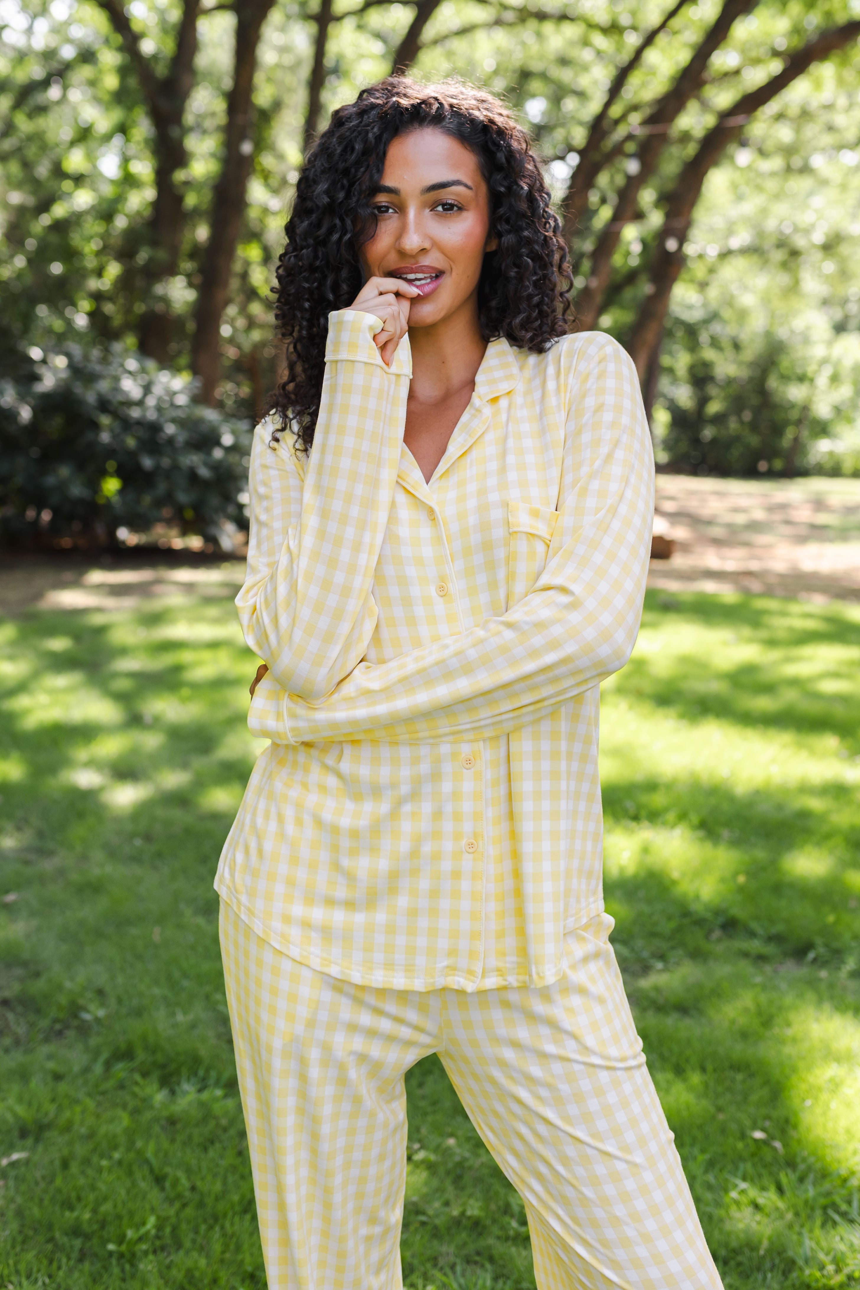 A woman wearing a light yellow and white gingham pajama set stands outdoors on a grassy lawn with trees in the background.