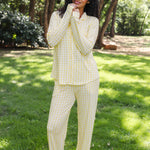 A woman wearing a light yellow and white gingham pajama set stands barefoot on a grassy lawn with trees in the background.