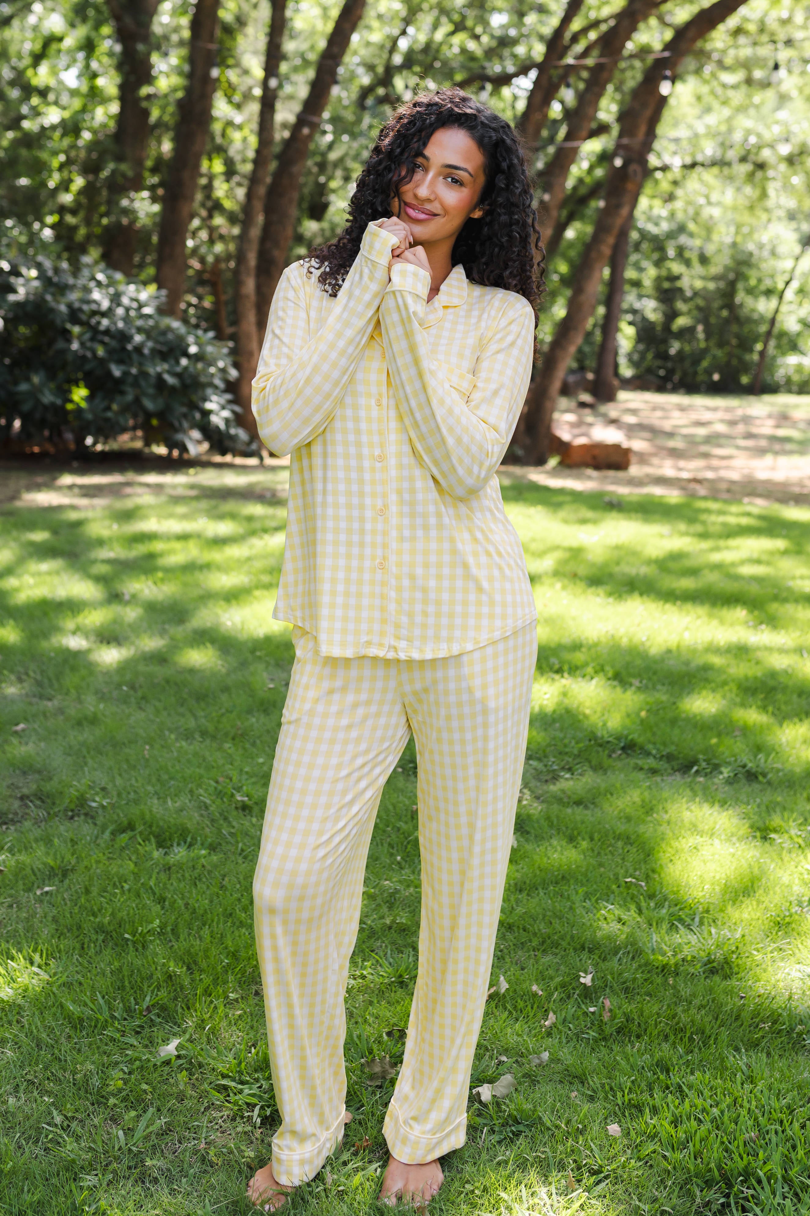 A woman wearing a light yellow and white gingham pajama set stands barefoot on a grassy lawn with trees in the background.