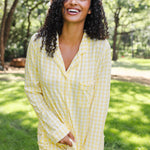 A woman with curly dark hair wearing a light yellow and white gingham pajama set, standing outdoors in a grassy park area with trees in the background.