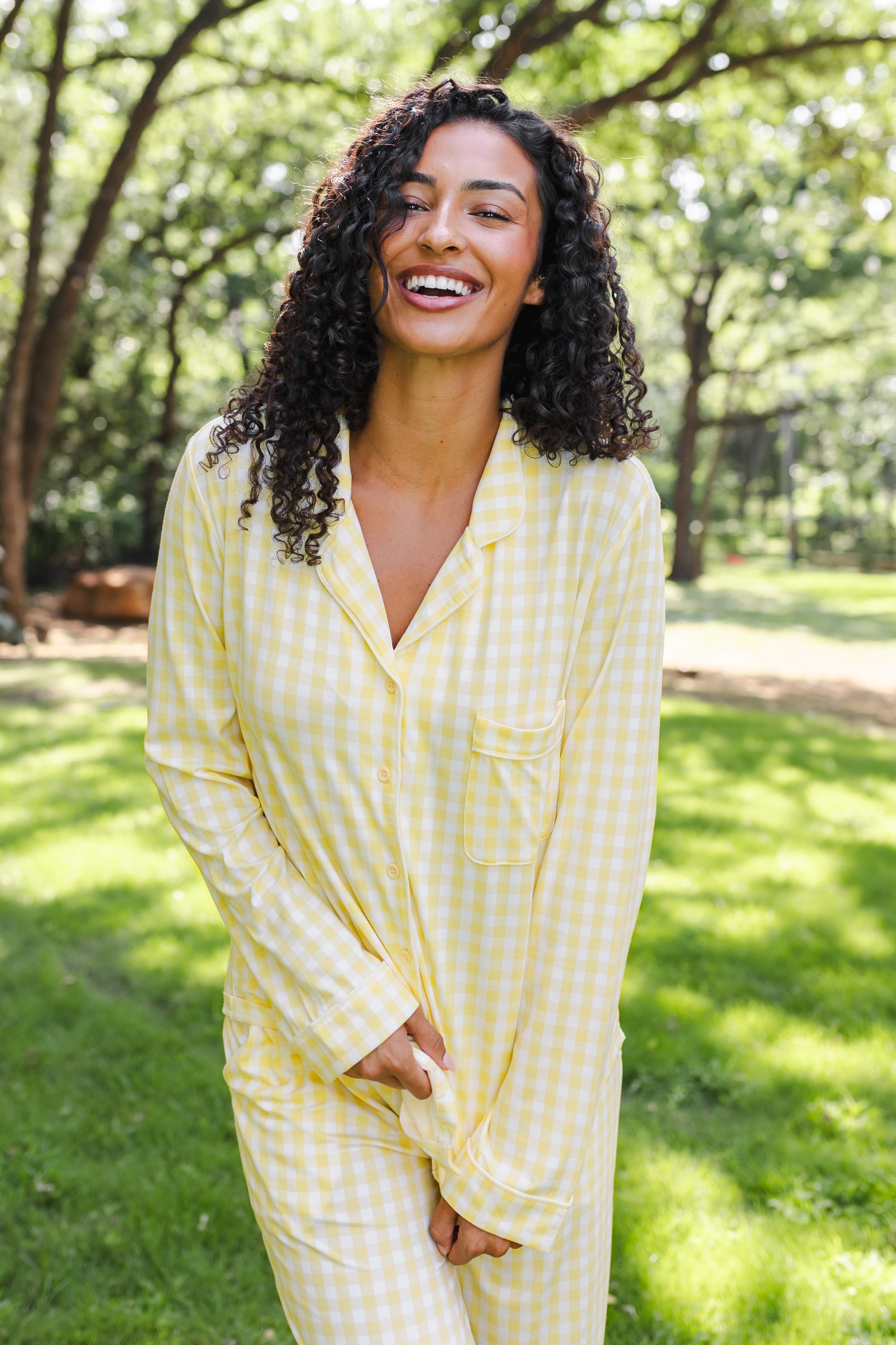 A woman with curly dark hair wearing a light yellow and white gingham pajama set, standing outdoors in a grassy park area with trees in the background.