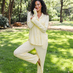 A woman wearing a light yellow and white checkered pajama set stands barefoot on a grassy lawn in a wooded park setting.