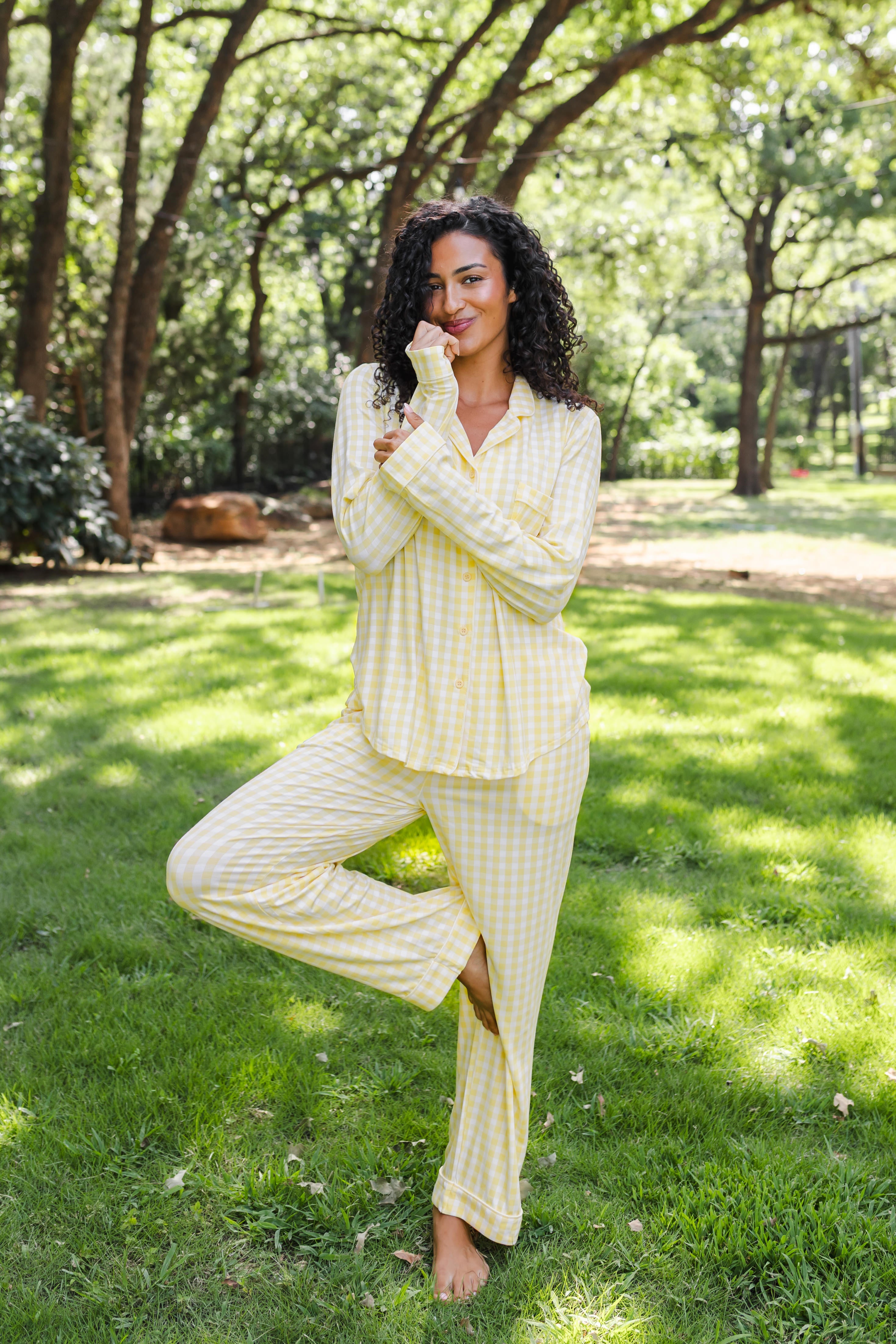 A woman wearing a light yellow and white checkered pajama set stands barefoot on a grassy lawn in a wooded park setting.