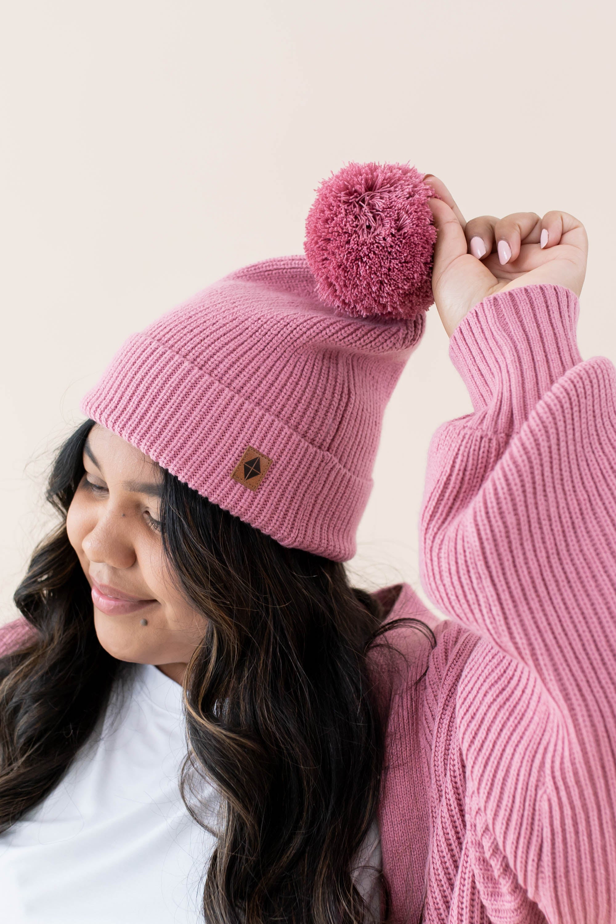 Focus is on the Pom on the hat and Kyte Baby's Leather Logo for the Chunky Knit Adult Pom Beanie in Apple Blossom 