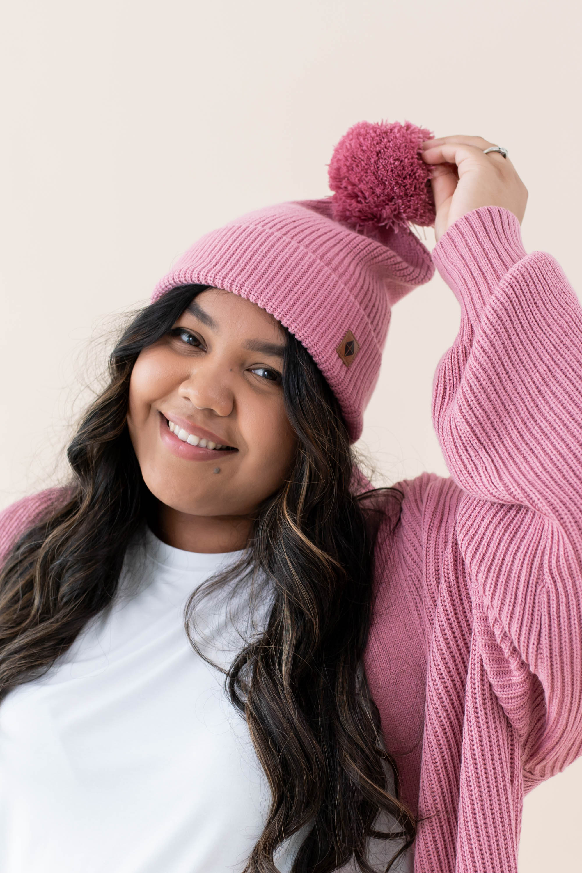 Smiling model wearing the Chunky Knit Adult Pom Beanie in Apple Blossom with one hand holding up the pom