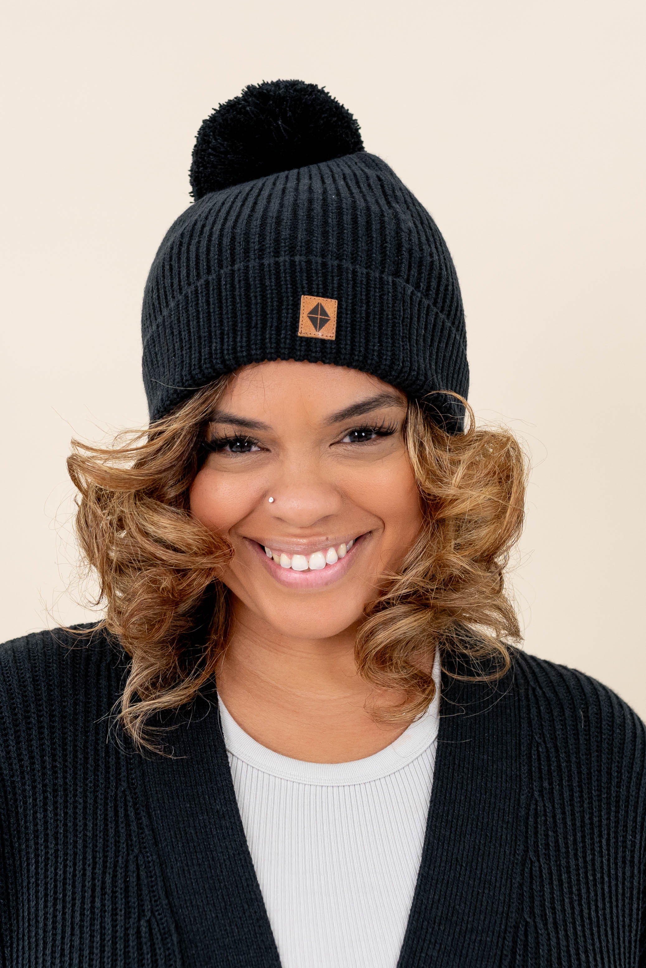 Smiling model wearing the Chunky Knit Adult Pom Beanie in Midnight with the Kyte Baby Leather Logo visible