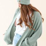 Model wearing the Chunky Knit Adult Pom Beanie in Thyme paired with a white shirt and Thyme Chunky Knit Oversized Women's Cardigan and jeans
