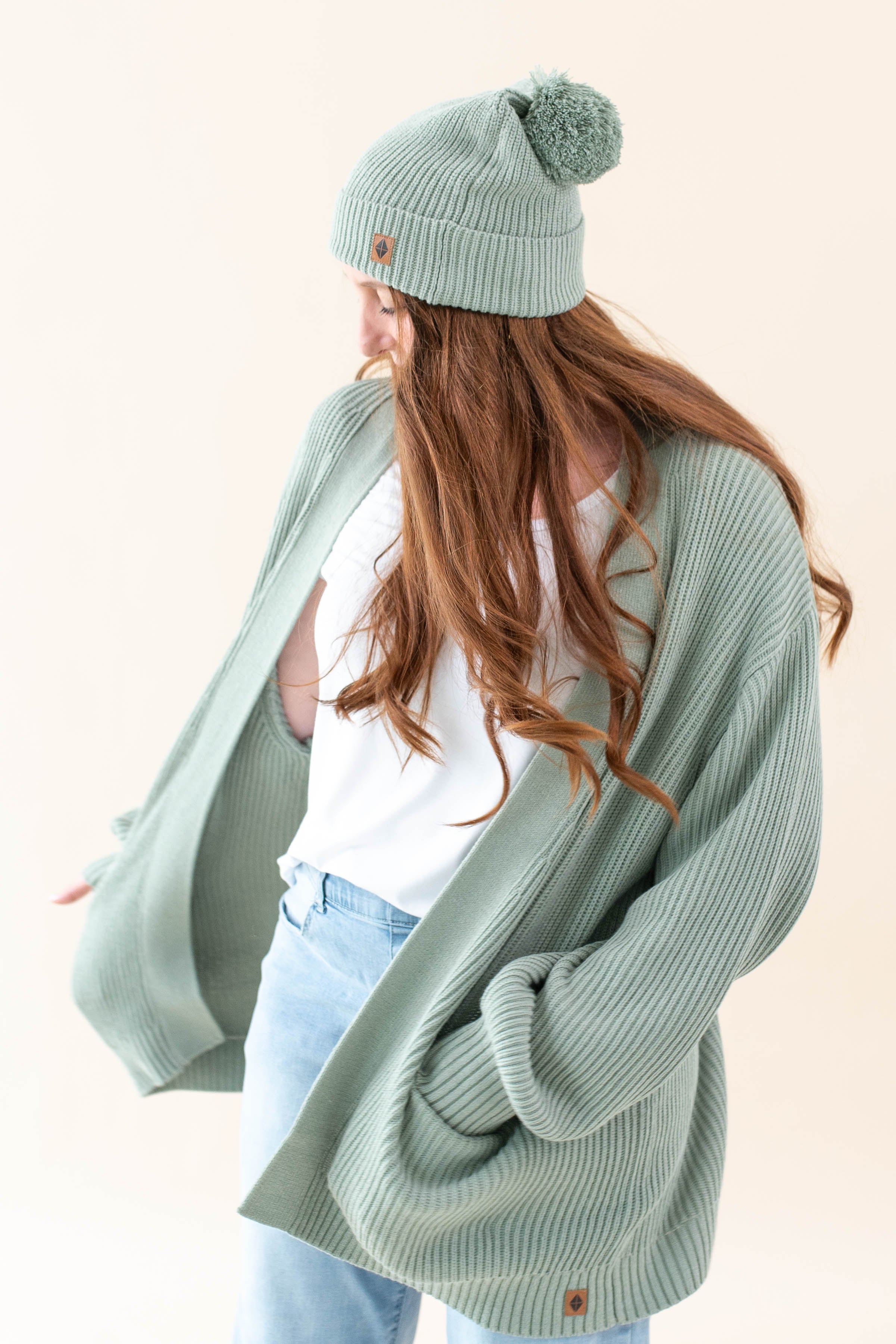 Model wearing the Chunky Knit Adult Pom Beanie in Thyme paired with a white shirt and Thyme Chunky Knit Oversized Women's Cardigan and jeans