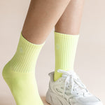 A close-up of a person's legs wearing neon yellow ribbed crew socks and a white chunky sneaker against a solid pale beige background.