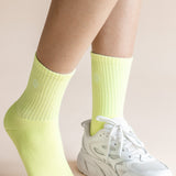 A close-up of a person's legs wearing neon yellow ribbed crew socks and a white chunky sneaker against a solid pale beige background.