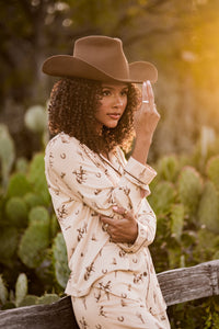 Female model wearing the Long-Sleeved Women's Pajama Set in Classic Cowboy and cowboy hat leaning up against a fence