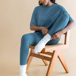 Woman sitting on a wooden chair wearing a teal outfit with white socks against a beige background