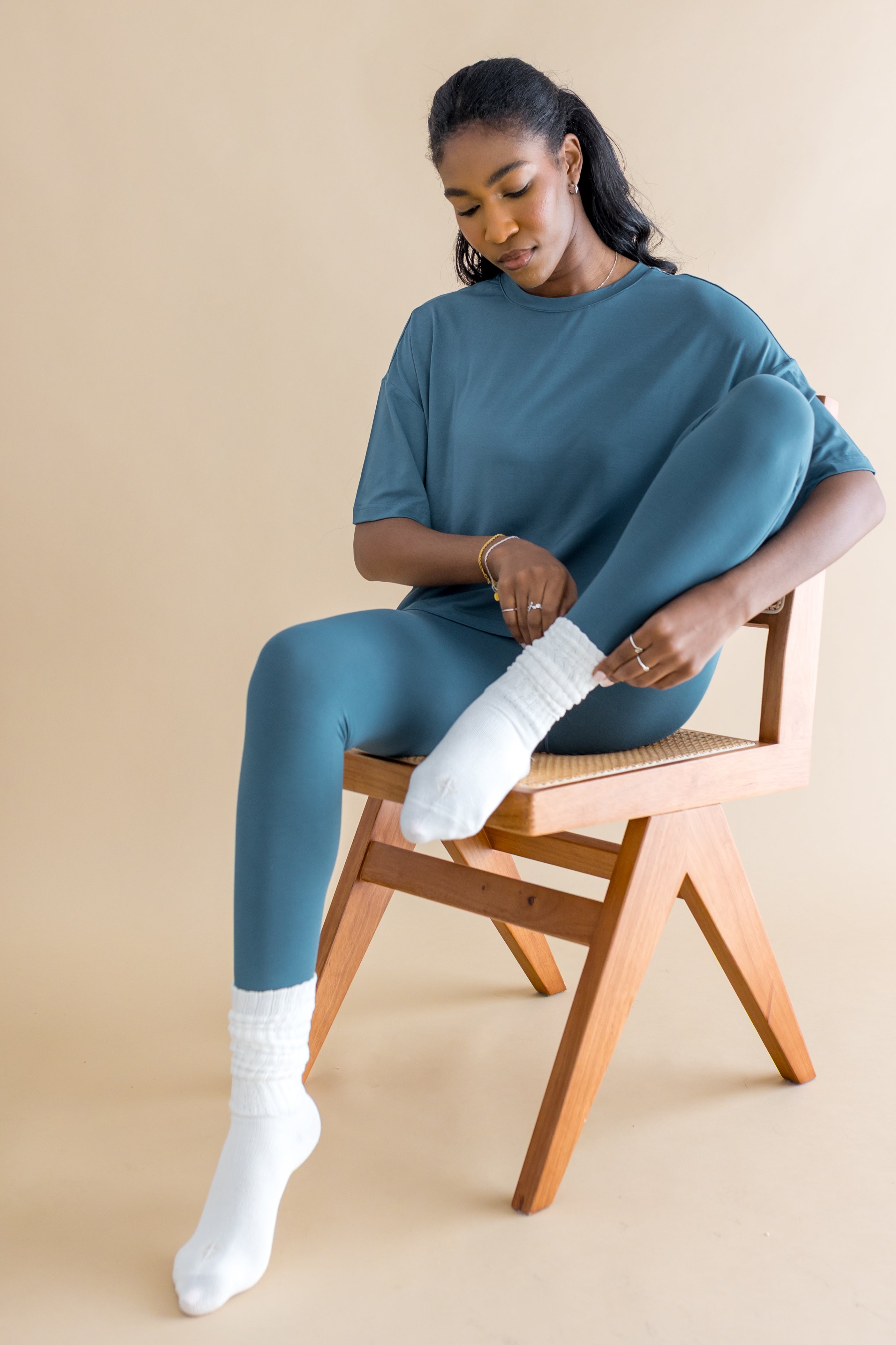 Woman sitting on a wooden chair wearing a teal outfit with white socks against a beige background