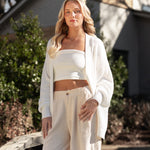Female model standing outside on a wooden deck wearing the Chunky Knit Women's Oversized Cardigan in Cloud overtop a white shirt and off white shorts